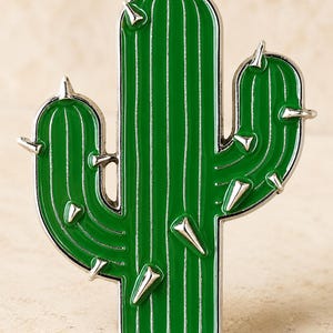 Peut inclure: Une broche en émail vert en forme de cactus saguaro avec un contour en métal argenté et des pointes argentées dépassant des côtés et de l'avant. Le cactus présente des lignes verticales en relief le long du corps principal et des deux bras.