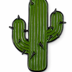 Könnte beinhalten: Eine grüne Emaille-Anstecknadel in Form eines Saguaro-Kaktus mit schwarzen Metallumrandungen und schwarzen Dornen. Der Kaktus besteht aus einer zentralen vertikalen Säule mit zwei gebogenen Seitenarmen, die mit vertikalen Rillen versehen sind.