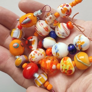 May include: A collection of colorful glass beads in various shapes and sizes, including round, oval, and irregular forms. The beads feature orange, white, red, and blue hues with speckled and swirled patterns. Some beads are strung together on orange cords.