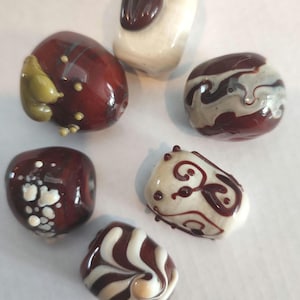 May include: A collection of artisan glass beads in various shapes and designs. The beads feature a mix of colors, including deep red, cream, and brown, with decorative swirls, dots, and patterns. Some beads have a glossy finish, while others have a matte appearance.