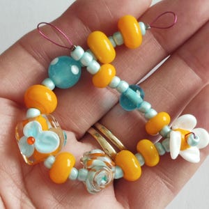 May include: A handmade bracelet featuring a variety of colorful glass beads. The bracelet includes orange, yellow, and light blue beads, with a flower and a polka-dot bead. The bracelet is held in a hand.