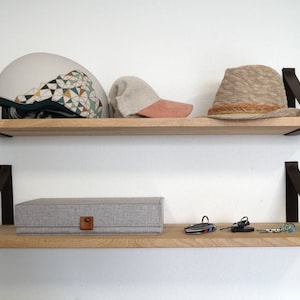 Op de afbeelding: Twee houten planken met zwarte leren banden, die een witte helm met geometrische patronen, een beige baseballpet met een roze klep, een beige geweven hoed, een grijze stoffen doos en sleutels bevatten.
