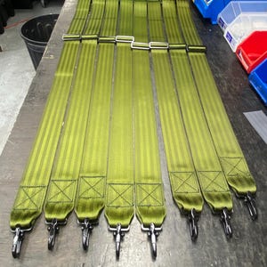 Może przedstawiać: Kolekcja oliwkowych pasków z metalowymi sprzączkami w kolorze srebrnym. Każdy pasek ma wzór w paski i metalowe zapięcie na końcu. Paski ułożone są na ciemnej powierzchni.