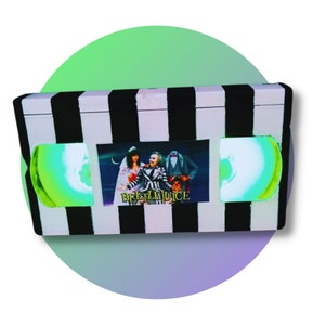 Puede incluir: Una caja a rayas blancas y negras con un póster de la película Beetlejuice. La caja tiene un póster rectangular de la película y dos carretes de plástico teñidos de verde. El texto "Beetlejuice" es visible en el póster.
