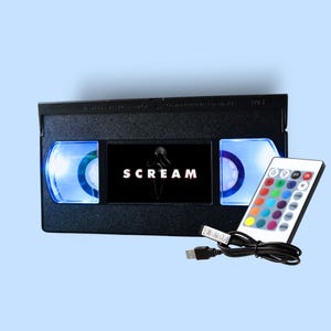 Puede incluir: Una cinta VHS negra con la palabra "SCREAM" en blanco, iluminada con luz azul. Se incluye un mando a distancia con múltiples opciones de color y un cable USB. Es un objeto decorativo.