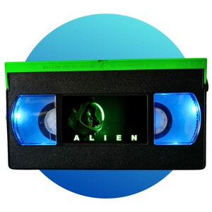May include: A black VHS tape with a green top and blue reels. The tape features the movie title "ALIEN" in white text and a green image of an astronaut. The top of the tape reads "Insert this side into recorder" and "Do not touch the tape inside."