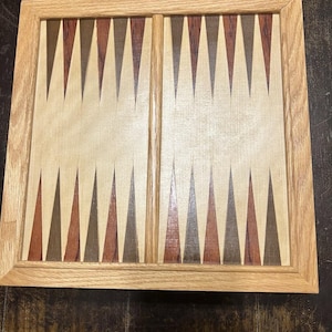 Pode incluir: Um tabuleiro de gamão quadrado de madeira com uma moldura de madeira clara e uma superfície de jogo decorada com pontos triangulares alternados em tons de castanho escuro, castanho avermelhado e bege claro, dispostos em duas secções separadas por um divisor central.