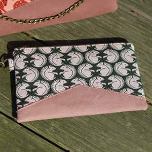 Könnte beinhalten: Eine kleine rosa und grüne Reißverschlusstasche mit einem Muster aus weißen Eichhörnchen auf dunkelgrünem Hintergrund. Die Tasche hat eine rosa Klappe mit einer spitzen Kante.