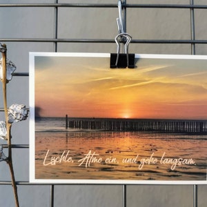 Könnte beinhalten: Eine Postkarte mit einem Sonnenuntergang über dem Meer, gehalten von einer schwarzen Klammer. Das Bild zeigt einen leuchtend orangefarbenen und gelben Himmel, der sich im Wasser spiegelt. Der Text unten lautet: "Lächle. Atme ein, und gehe langsam."