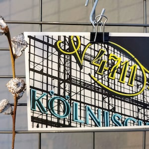 Könnte beinhalten: Eine Postkarte mit dem Text "4711 KÖLNISCH" in Gelb und Türkis vor einem schwarz-weißen architektonischen Hintergrund. Die Postkarte wird von einer schwarzen Klammer an einem Drahtgitter gehalten. Links ist eine getrocknete Pflanze mit weißen Akzenten.