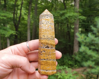 May include: A yellow and brown tiger's eye crystal point, also known as a wand, is held in a hand. The crystal has a pointed tip and a smooth, polished surface.
