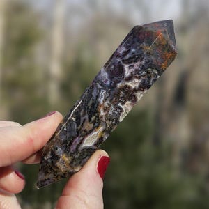 May include: A polished, elongated crystal point with a mix of colors including black, white, purple, and orange. The crystal is held in a hand with red nail polish. The crystal's surface has a unique, natural pattern.