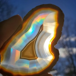 May include: A slice of agate with a rainbow iridescence. The agate is translucent and has a brown outer edge. The agate is held in a hand against a blue sky.