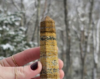 May include: A yellow, brown, and black banded crystal point, also known as a bumblebee jasper point, is held in a hand. The crystal is about 5 inches long.
