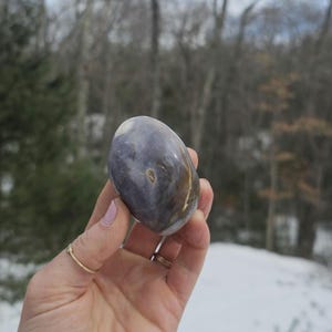 May include: A smooth, polished purple and brown agate stone with a swirling pattern. The stone is oval shaped and has a natural, organic look.
