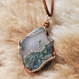 May include: A pendant necklace featuring a green and white agate stone wrapped in copper wire. The stone has a unique pattern and is set on a brown leather cord.