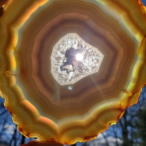 May include: A large, round, agate slice with a hole in the center. The agate is brown, orange, and white with iridescent rainbow colors. The sun is shining through the hole in the agate.