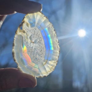 May include: A clear, iridescent agate slice with a rainbow sheen. The agate is held in a hand against a blue sky background.