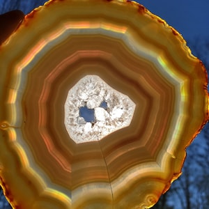 May include: A large, round, agate slice with a hole in the center. The agate is a mix of brown, white, and orange colors with iridescent rainbow hues. The agate is held up to the sun, and the light shines through the hole.