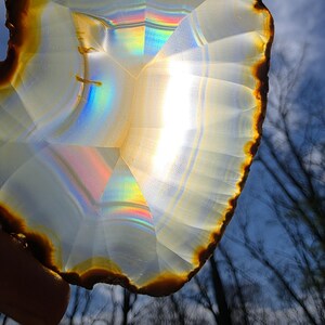 May include: A translucent agate slice with a brown and gold outer edge. The stone displays internal rainbow refractions and is held against a backdrop of a blue sky and tree silhouettes.