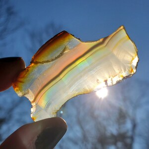 May include: A piece of iridescent agate with brown, orange, and white banding. The agate is held up to the sun, showing off its rainbow colors.