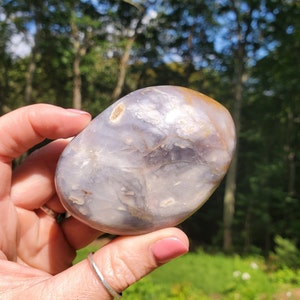 May include: A smooth, oval-shaped, light purple agate stone with brown and white markings. The stone is being held in a hand.
