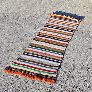 May include: A long, striped rug with orange, brown, white, blue, and green stripes. The rug has fringe on the ends and is lying on a gray asphalt surface.