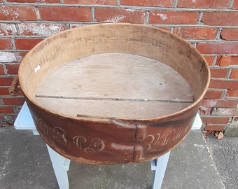 May include: A round wooden container with a brown leather rim and a white inscription. The container is sitting on a white stool against a red brick wall.