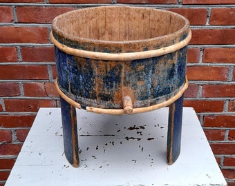 May include: A wooden bucket with blue and yellow paint, sitting on a white table with three legs. The bucket has a round top and a flat bottom. The bucket is made of wood and has a rustic look.