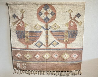 May include: A woven wall hanging with a beige background and a brown, blue and white geometric design. The design features two stylised birds flanking a central tree with a circular top. The bottom border is brown with a repeating pattern of white and blue flowers. The hanging has a fringe border and the initials "ILS" are woven into the bottom corner.