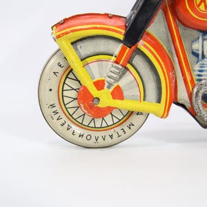 May include: Vintage toy motorcycle in red, yellow, and white. The front wheel has a white tire with black spokes and text. The motorcycle has a blue rider and a silver exhaust pipe. The toy is made of metal.