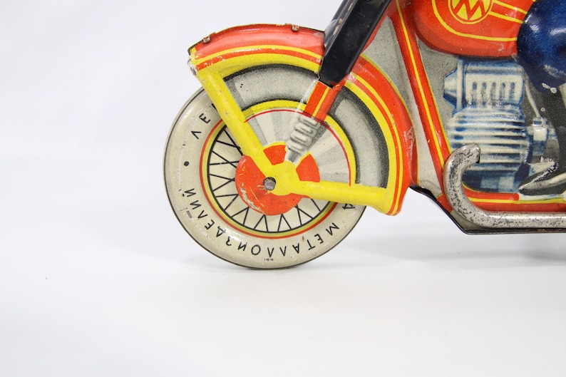 May include: Vintage toy motorcycle in red, yellow, and white. The front wheel has a white tire with black spokes and text. The motorcycle has a blue rider and a silver exhaust pipe. The toy is made of metal.