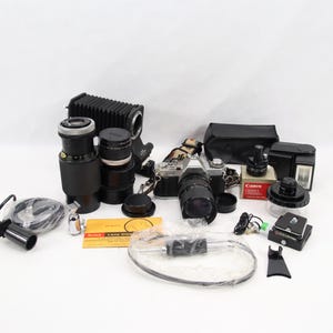 Peut inclure: Ensemble de matériel photographique comprenant un boîtier d'appareil photo, des objectifs, un flash et divers accessoires. Les objectifs sont noirs et le boîtier de l'appareil photo est argenté et noir. Les articles sont disposés sur une surface blanche.