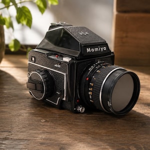 May include: A black Mamiya M645 medium format film camera with a lens attached. The camera is on a wooden surface. The lens has a filter and the camera body has the name "Mamiya" printed on it.