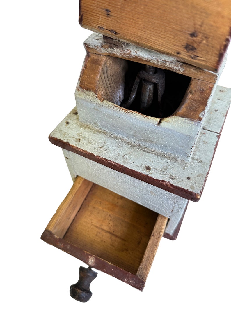 May include: Antique wooden coffee grinder with a drawer and a metal grinding mechanism. The grinder is painted white with red accents and has a wooden handle.
