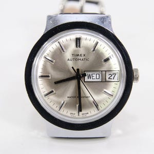 Könnte beinhalten: Silberfarbene Timex Automatic-Uhr mit schwarzer Lünette und hellem Zifferblatt. Die Uhr zeigt Tag und Datum an, wobei der Tag "WED" und das Datum "27" lautet. Die Uhr hat ein silbernes Armband.