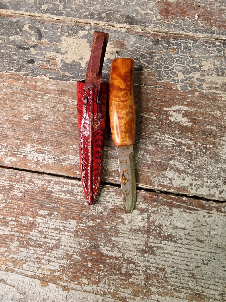 Puede incluir: Un peque&ntilde;o cuchillo con mango de madera y una funda de cuero. El cuchillo tiene una hoja lisa y la funda es roja con un patr&oacute;n decorativo.