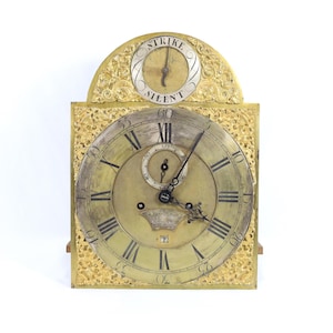 May include: Antique gold-toned clock with Roman numerals and ornate detailing. The clock face features a smaller dial at the top with the words "STRIKE" and "SILENT". The clock's design includes intricate patterns and a square shape.