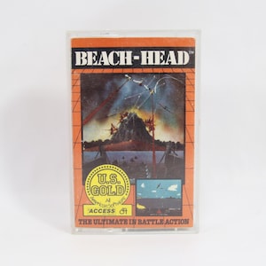 Puede incluir: Un cartucho de videojuego vintage de "Beach-Head" en una funda de plástico transparente. La etiqueta naranja y negra presenta una ilustración dramática de una escena de batalla con el texto "U.S. GOLD" y "THE ULTIMATE IN BATTLE ACTION".
