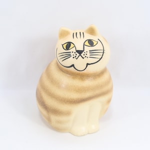 May include: A ceramic cat figurine with a cream-colored body and brown stripes. The cat has yellow eyes, black whiskers, and a smiling face. The figurine is sitting upright with its legs tucked beneath it.
