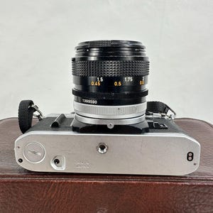May include: A silver Canon camera with a black lens. The lens has markings for 0.45, 0.5, 1.5, 1.75, and 2. The camera has a black strap and is sitting on a brown leather case.