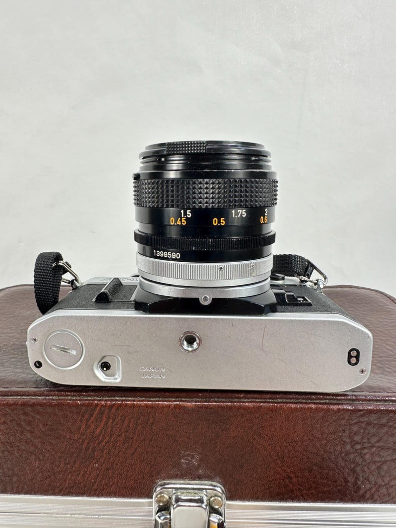 May include: A silver Canon camera with a black lens. The lens has markings for 0.45, 0.5, 1.5, 1.75, and 2. The camera has a black strap and is sitting on a brown leather case.