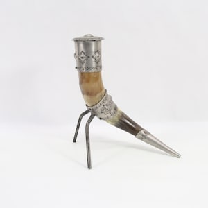 Op de afbeelding: Een decoratieve drinkhoorn met een metalen rand en punt, ondersteund door een metalen driepoot. De hoorn is lichtbruin van kleur met donkerbruine accenten en zilverkleurige metalen details.