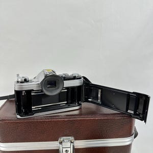 May include: A silver 35mm film camera with a black lens and a black film back. The camera is sitting on top of a brown leather carrying case with a silver latch. Two additional lenses are visible in the bottom of the image.