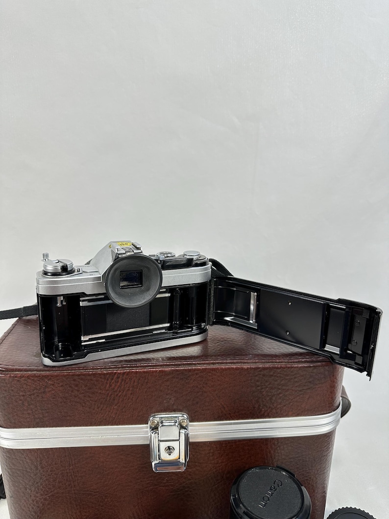 May include: A silver 35mm film camera with a black lens and a black film back. The camera is sitting on top of a brown leather carrying case with a silver latch. Two additional lenses are visible in the bottom of the image.