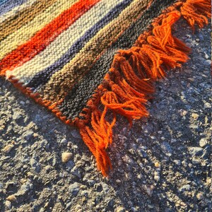 May include: A woven rug with orange, brown, white, and blue stripes. The rug has a fringe on one edge.