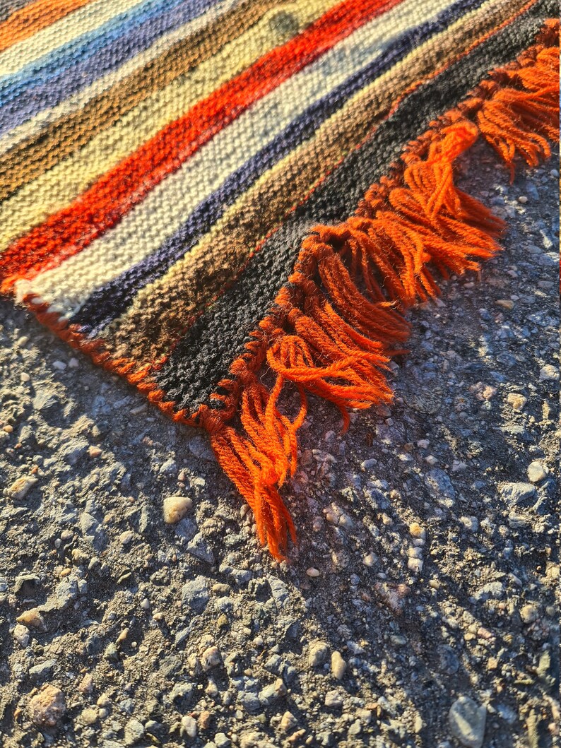 May include: A woven rug with orange, brown, white, and blue stripes. The rug has a fringe on one edge.