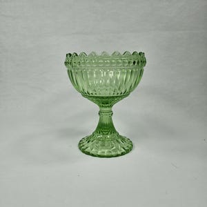 May include: A green glass pedestal bowl with a scalloped rim and a textured design. The bowl is on a white background.
