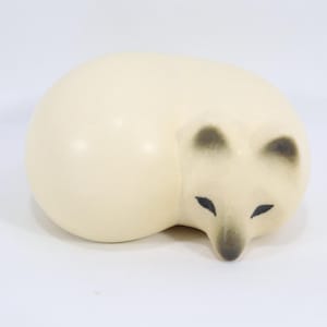 May include: A cream-colored ceramic cat figurine in a curled-up position. The cat has black markings on its ears and nose, with black, almond-shaped eyes. The figurine is on a white background.