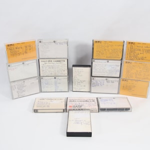 以下が含まれることがあります： 手書きのラベルが付いた、いくつかのヴィンテージオーディオカセットのコレクション。テープは透明なプラスチックケースに入っており、一部は黄ばんだ紙のインサートが付いています。画像は白い背景にあります。